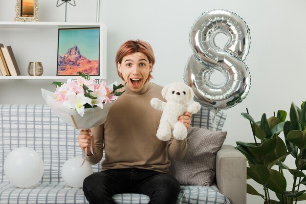 Hübscher Kerl am glücklichen Frauentag, der Blumenstrauß mit Teddybär hält, der auf Sofa im Wohnzimmer sitzt