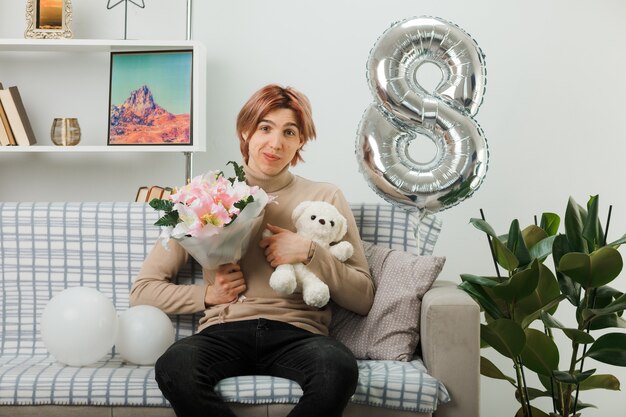 Hübscher Kerl am glücklichen Frauentag, der Blumenstrauß mit Teddybär hält, der auf Sofa im Wohnzimmer sitzt