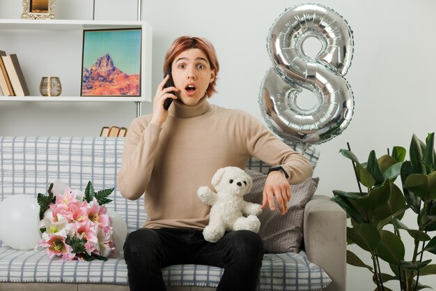 Hübscher Kerl am glücklichen Frauentag, der Teddybären hält, spricht am Telefon, das auf Sofa im Wohnzimmer sitzt