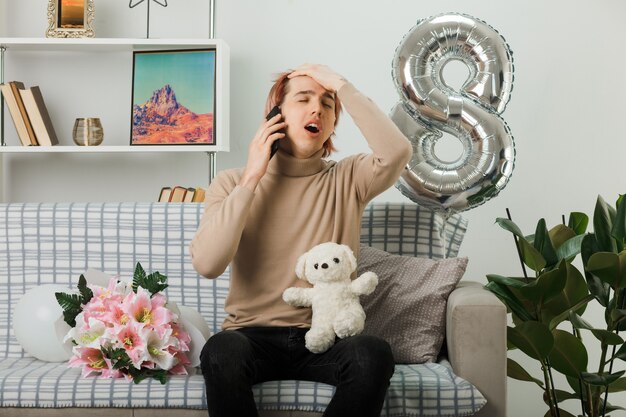 Hübscher Kerl am glücklichen Frauentag, der Teddybären hält, spricht am Telefon, das auf Sofa im Wohnzimmer sitzt