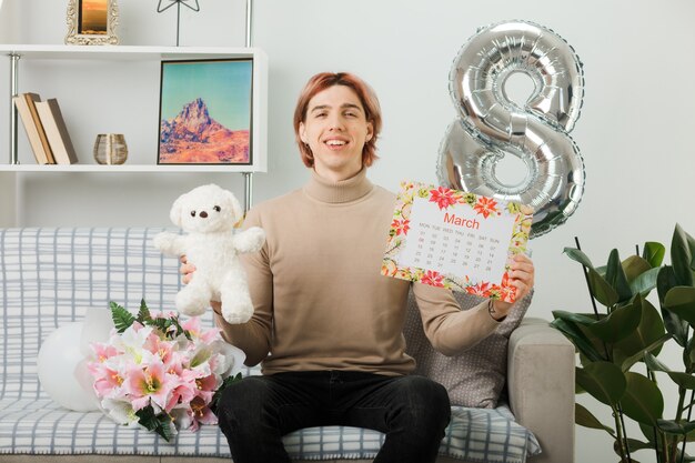 Hübscher Kerl am glücklichen Frauentag, der Teddybären mit Kalender hält, der auf Sofa im Wohnzimmer sitzt