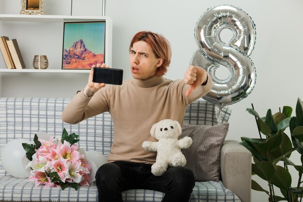 Hübscher Kerl am glücklichen Frauentag, der Teddybären mit Telefon hält, der auf Sofa im Wohnzimmer sitzt