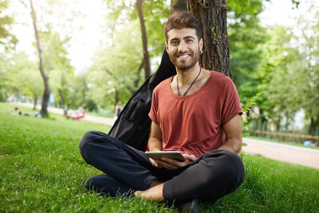 Hübscher Kerl lehnt sich an Baum, liest E-Buch mit digitalem Tablett im Park