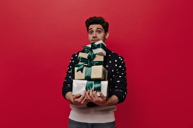 Hübscher kleiner Junge mit brünetten Haaren und Weihnachtstrikot, der einen Stapel Geschenkboxen hält, die gerade schauen und isoliert auf rotem Hintergrund posieren