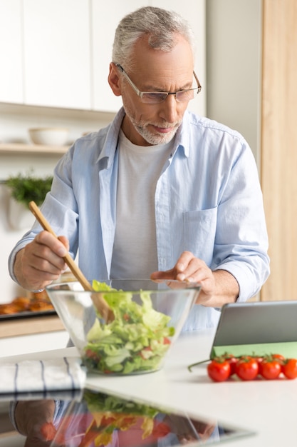 Hübscher konzentrierter reifer Mann, der Salat mit Tablette kocht