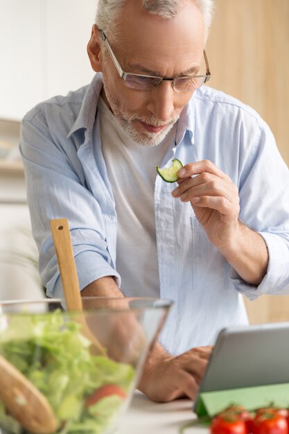 Hübscher konzentrierter reifer Mann, der Salat mit Tablette kocht