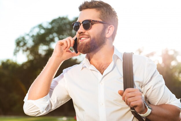 Hübscher lächelnder bärtiger Mann in der Sonnenbrille, die durch das Smartphone draußen spricht und wegschaut