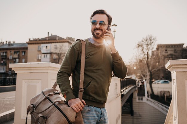 Hübscher lächelnder stilvoller Hipster-Mann, der in der Stadtstraße mit Leder spricht, das am Telefon auf Geschäftsreisetasche trägt, die Sweatshot und Sonnenbrille, Stadtstil-Trend, sonniger Tag, Reisen trägt