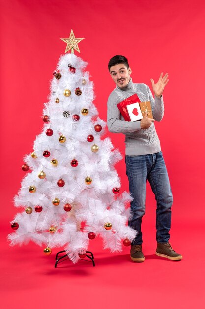 Hübscher lustiger junger Mann, der nahe dem verzierten weißen Neujahrsbaum steht und seine Geschenke hält