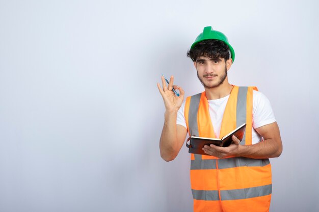 Hübscher männlicher Ingenieur im grünen Helm mit Notizbuch, das über weißer Wand steht. Hochwertiges Foto