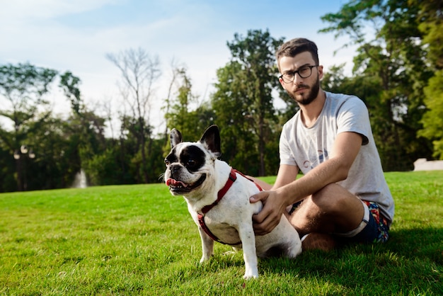 Hübscher Mann, der mit französischer Bulldogge auf Gras im Park sitzt