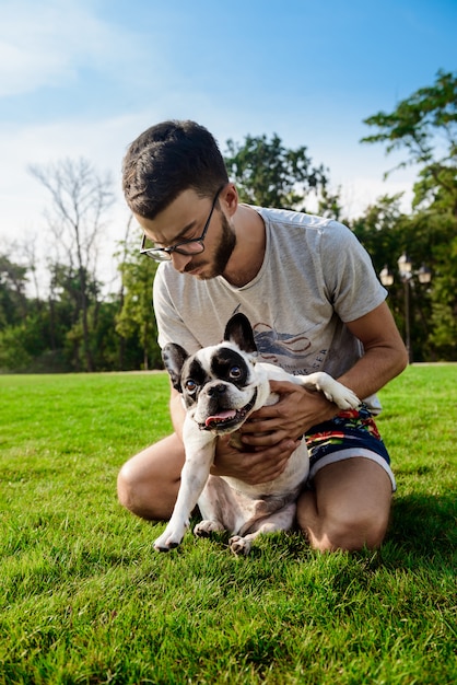 Hübscher Mann, der mit französischer Bulldogge auf Gras im Park sitzt