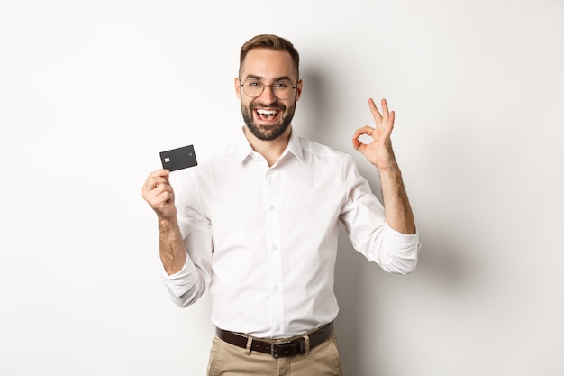 Hübscher Mann, der seine Kreditkarte und das OK-Zeichen zeigt, Bank empfiehlt, stehenden Kopierraum