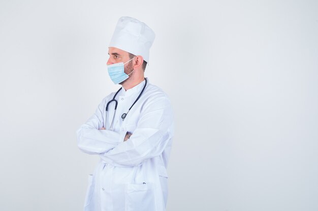Hübscher Mann, der Stethoskop um Hals hält, stehende Arme verschränkt im weißen medizinischen Laborkittel, Maske und selbstbewusst aussehend. Vorderansicht.