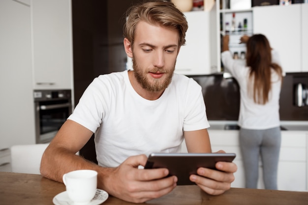 Hübscher Mann, der Tablette betrachtet. Freundin in der Küche