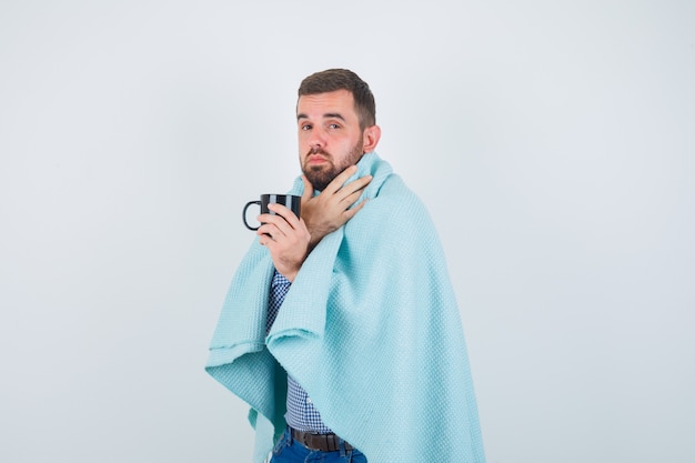 Hübscher Mann, der Tasse Tee hält, Halsschmerzen in Hemd, Jeans, Schal hat und erschöpft aussieht, Vorderansicht.