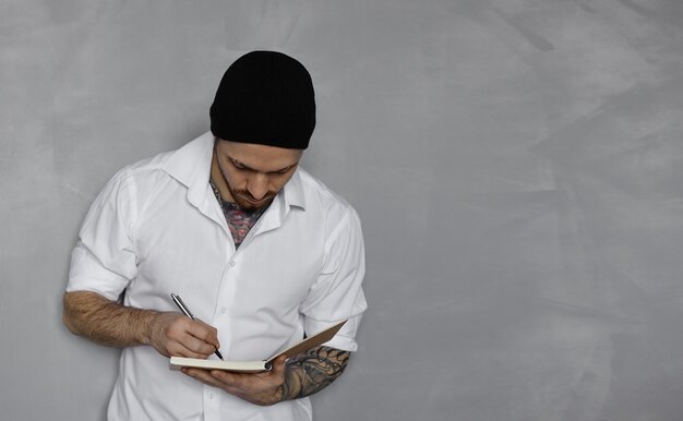 Hübscher Mann im weißen Hemd und im schwarzen Hut bleiben nahe grauer Wand und schreiben Notizen in Notizblock