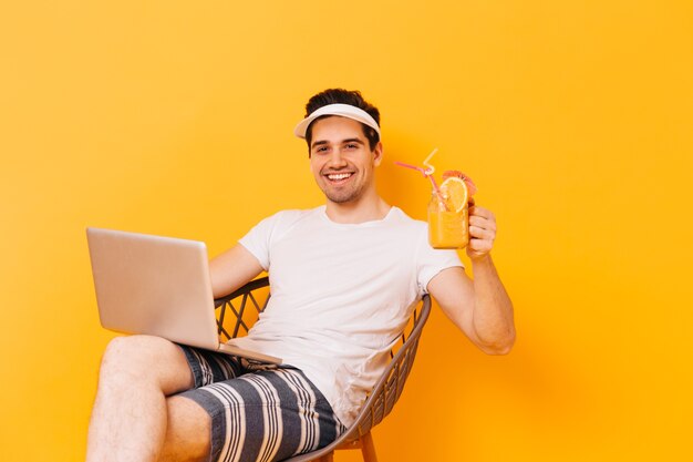 Hübscher Mann mit braunen Augen im Strandoutfit hob ein Glas Saft und lächelte, während er im Laptop arbeitete.