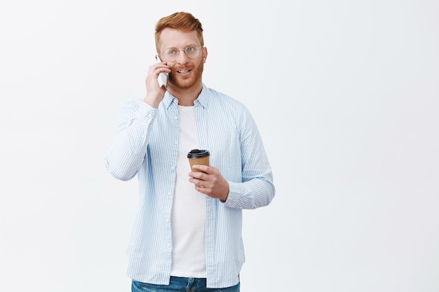 Hübscher moderner geschäftsmann mit roten haaren in gläsern, tasse des getränks haltend und über smartphone lässig über graue wand sprechend