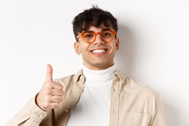Hübscher moderner Typ in Brille und Ohrring, zufrieden lächelnd und Daumen hoch, empfehlen Unternehmen, stehend auf weißem Hintergrund