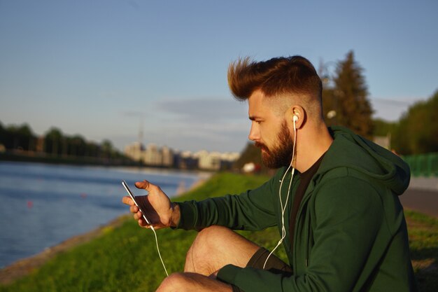 Hübscher modischer junger Mann mit stilvoller Frisur und dickem Bart, der friedlichen Sommermorgen draußen genießt, am See sitzt und Musiktitel unter Verwendung der Online-App auf seinem Handy hört