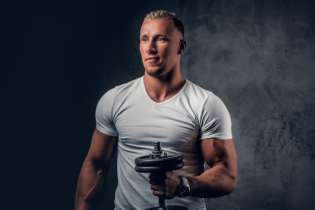 Kostenloses Foto hübscher muskulöser blonder mann in einem weißen t-shirt hält hantel über grauem künstlerischem hintergrund.