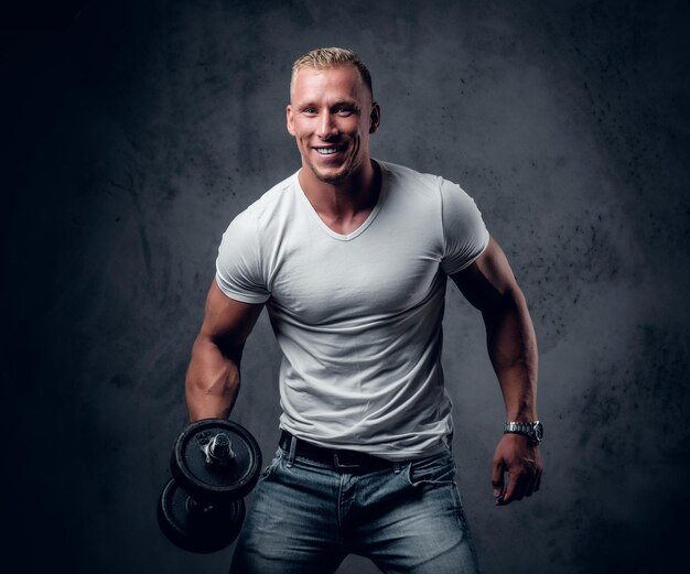 Hübscher muskulöser blonder Mann in einem weißen T-Shirt hält Hantel über grauem künstlerischem Hintergrund.