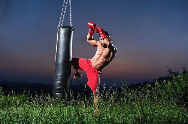 Hübscher muskulöser junger Kickboxer mit nacktem Oberkörper, der mit einem Boxsack im Freien trainiert.