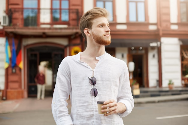 Hübscher rothaariger bärtiger Mann mit stilvollem Haarschnitt im weißen Hemd, das durch Stadt geht und Kaffee am Morgen vor hartem Arbeitstag trinkt.