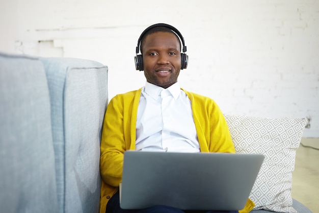 Hübscher schwarzer männlicher Student, der gelbe Strickjacke über weißem Hemd trägt, das zu Hause studiert, Laptop und Kopfhörer verwendet, Vortrag online hört. Glücklicher Mann, der Musik über Headset auf Sofa genießt