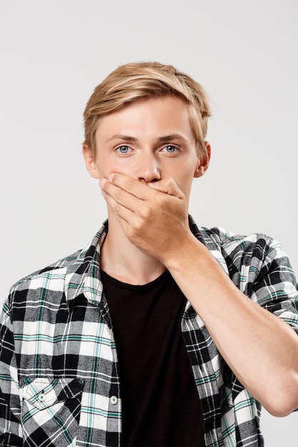 hübscher selbstbewusster blonder junger Mann, der lässiges kariertes Hemd trägt, das Mund mit Hand bedeckt, auf grauer Wand