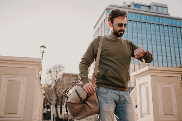 Hübscher stilvoller bärtiger Mann, der in Stadtstraße mit Lederreisetasche trägt, die Sweatshirt und Sonnenbrille, Stadtarttrend, sonniger Tag trägt, auf Uhr schaut