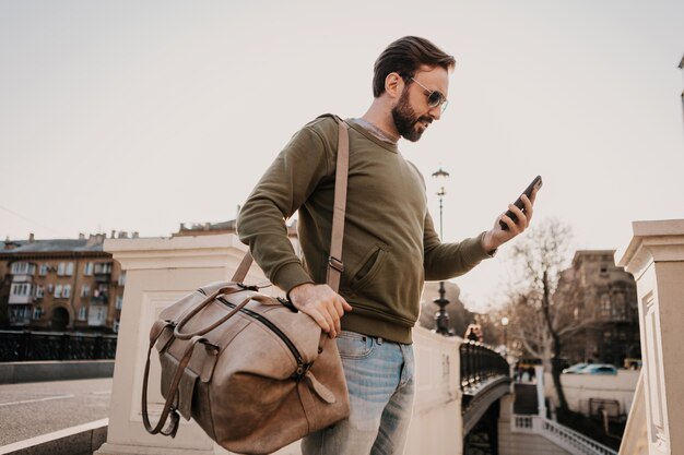 Hübscher stilvoller Hipster-Mann, der in der Stadtstraße mit Ledertasche unter Verwendung der Telefonnavigationsanwendung geht, Reise tragendes Sweatshirt und Sonnenbrille, Stadtstil-Trend