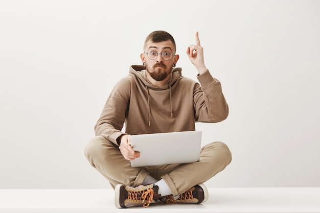 Hübscher Student sitzt mit Laptop auf gekreuzten Beinen und hebt den Zeigefinger, hat eine gute Idee