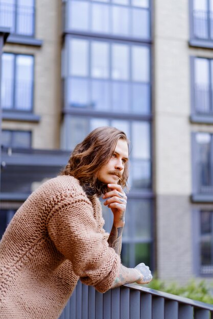 hübscher tätowierter sexy Mann mit langen Haaren und Schnurrbart in einem Strickpullover. Porträt außerhalb Nahaufnahme.