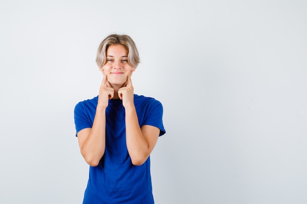 Hübscher Teenager, der die Finger auf den Wangen im blauen T-Shirt hält und fröhlich aussieht, Vorderansicht.