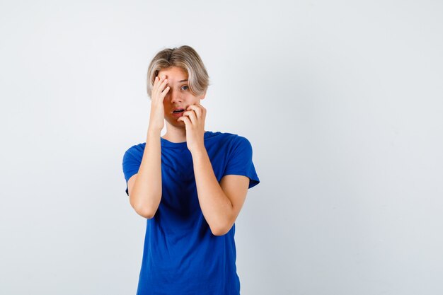 Hübscher Teenager, der die Hände im blauen T-Shirt auf dem Gesicht hält und verängstigt aussieht, Vorderansicht.