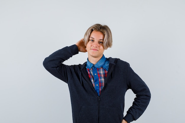 Hübscher teenager, der die hand hinter dem kopf in hemd, hoodie hält und fröhlich aussieht. vorderansicht.