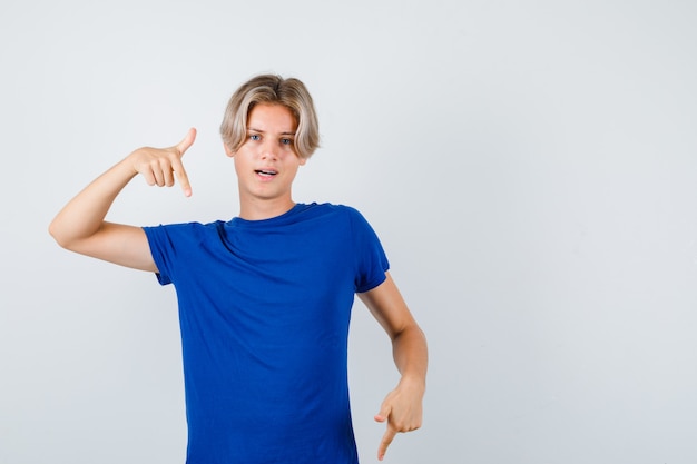 Hübscher Teenager, der im blauen T-Shirt nach unten zeigt und selbstbewusst aussieht, Vorderansicht.