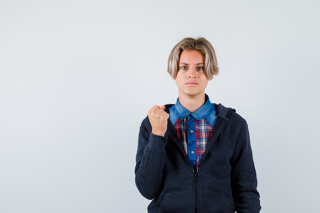 Hübscher Teenager im Hemd, Hoodie, der geballte Faust zeigt und wütend aussieht, Vorderansicht.