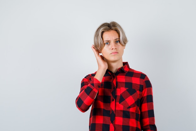 Hübscher Teenager im karierten Hemd mit der Hand in der Nähe des Ohrs und verwirrt, Vorderansicht.