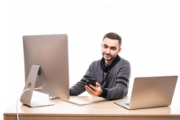 Hübscher Unternehmer, der hinter seinem Schreibtisch mit Laptop und PC sitzt und auf Handy lokalisiert auf Weiß tippt