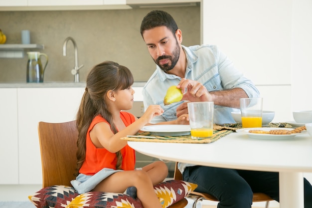 Hübscher Vater, der seinem Mädchen Obst gibt, während sie zusammen in der Küche frühstücken
