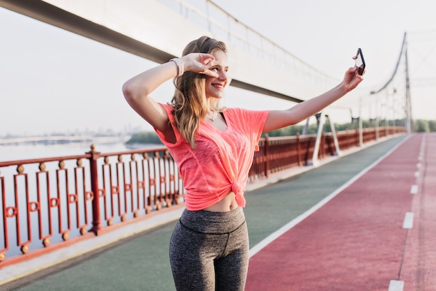 Hübscher weiblicher Läufer, der Smartphone für Selfie an Aschenbahn verwendet. Sinnliches Mädchen in Sportkleidung, das Foto von sich selbst macht.
