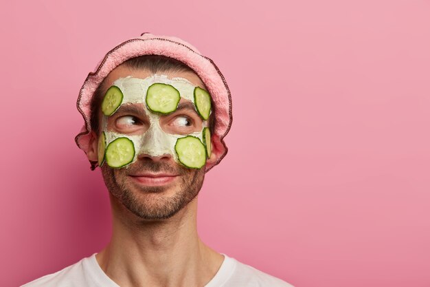 Hübscher zufriedener Mann bekommt Gesichtsbehandlung, trägt Tonmaske mit Gurkenscheiben auf, genießt Schönheitsbehandlungen, hat Borsten