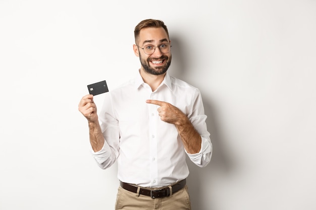 Hübscher zufriedener Mann in den Gläsern, die auf Kreditkarte zeigen, zufrieden mit Bankdienstleistungen, stehend