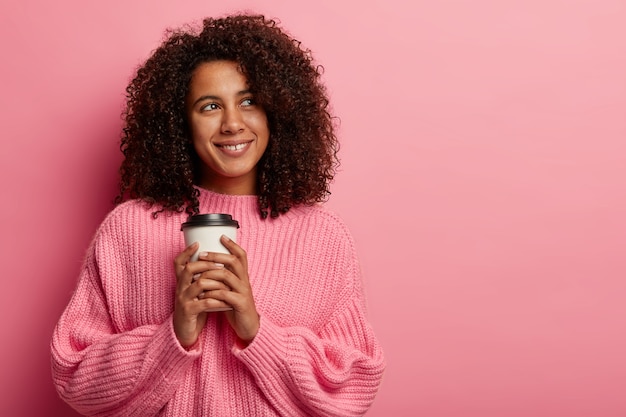 Hübsches Afro-Mädchen hält eine Kaffeetasse aus Papier, genießt die Freizeit, schaut mit einem Lächeln beiseite und trägt einen übergroßen Pullover