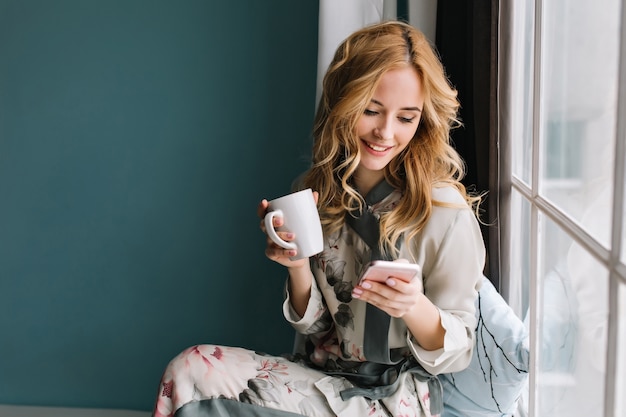 Hübsches blondes Mädchen, das auf Fensterbank mit Tasse Kaffee, Tee und Smartphone in den Händen sitzt. Sie hat langes blondes, welliges Haar, lächelt und schaut auf ihr Handy. Trage einen schönen Seidenpyjama.