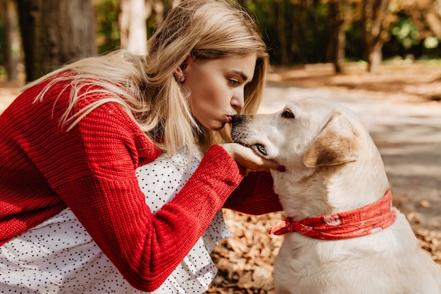 Hübsches blondes Mädchen, das ihren schönen Labradour küsst. Junge Frau, die rote Farbe mit ihrem Hund im Herbstpark trägt.