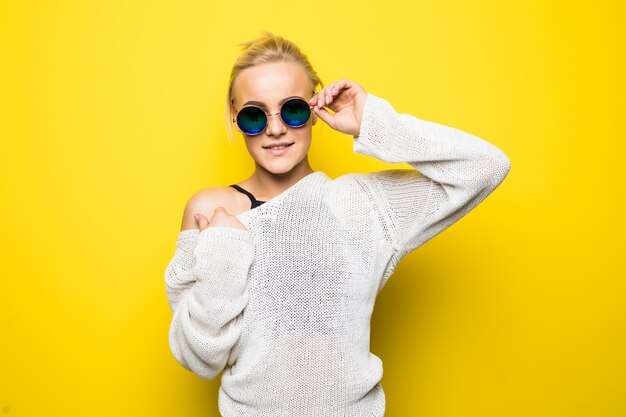 Hübsches blondes Mädchen im modernen weißen Pullover in der strahlend blauen Sonnenbrille posiert auf Gelb